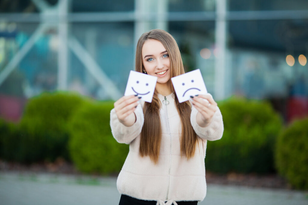 A smiling woman holding two cards—one with a happy face and one with a sad face—symbolizing the role of emotion in marketing.