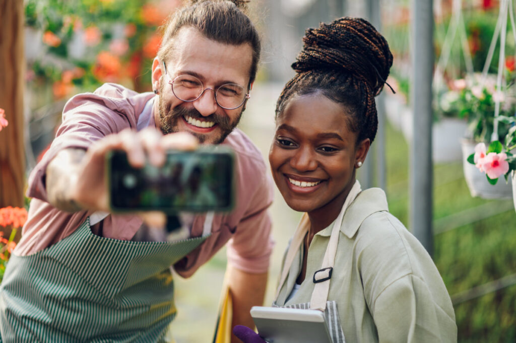 Two happy gardeners taking a selfie in a greenhouse, showcasing authenticity on social media through genuine connections.