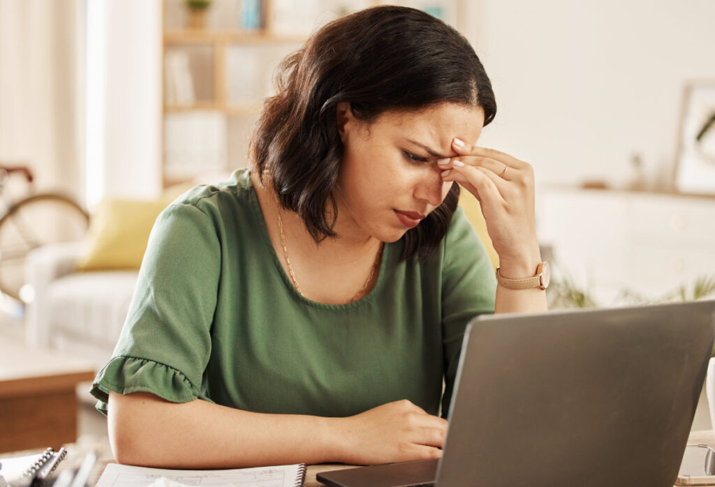 A frustrated woman sitting at a desk, holding her forehead while looking at a laptop, struggling with writing about herself.