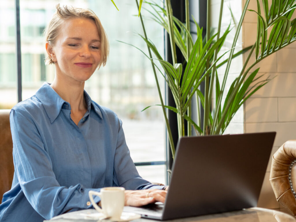 A smiling woman typing on a laptop in a café, representing business blogging and the importance of avoiding common blogging mistakes.