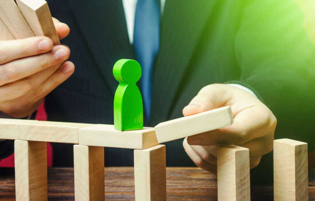 A businessman in a suit placing wooden blocks to create a bridge for a green figure, symbolizing overcoming marketing roadblocks.