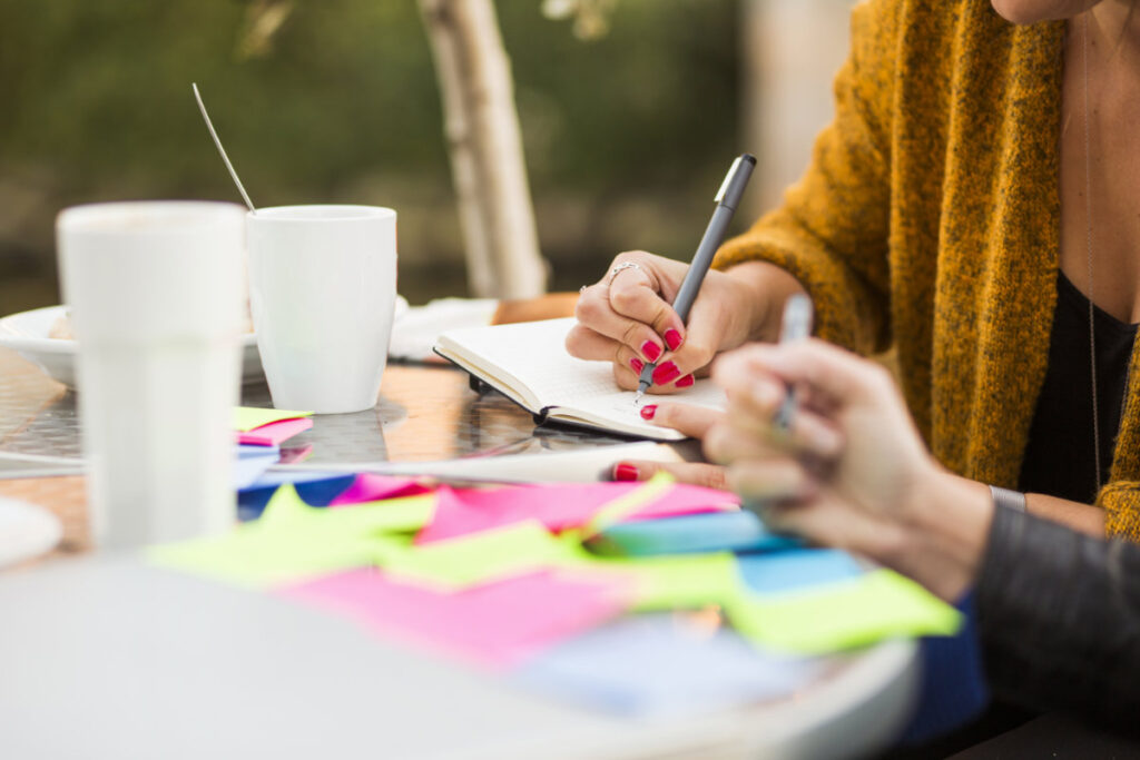 Two people writing in a notebook and organizing colorful sticky notes during a brainstorming session, focusing on setting attainable marketing goals.